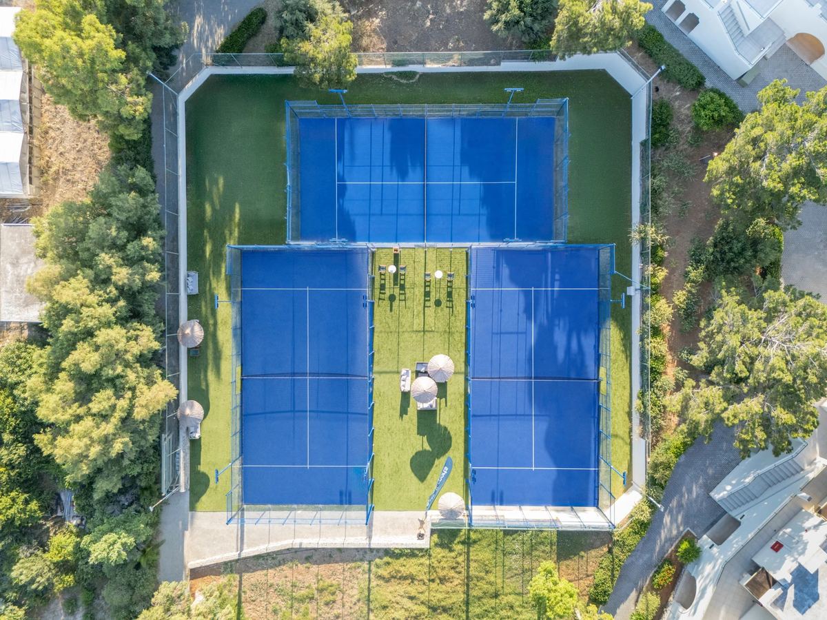 Luftaufnahme von drei blauen Padelplätzen mit Glaswänden, dazwischen eine Rasenfläche mit Sonnenschirmen