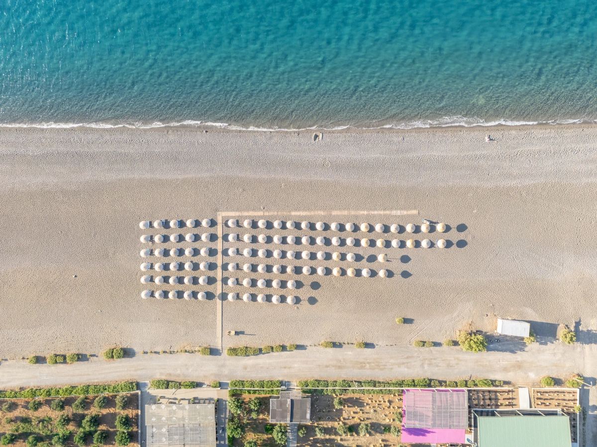 Strand vor dem Almyra senkrecht von oben: türkisfarbenes Meer, grauer Sand und Reihen von Sonnenschirmen
